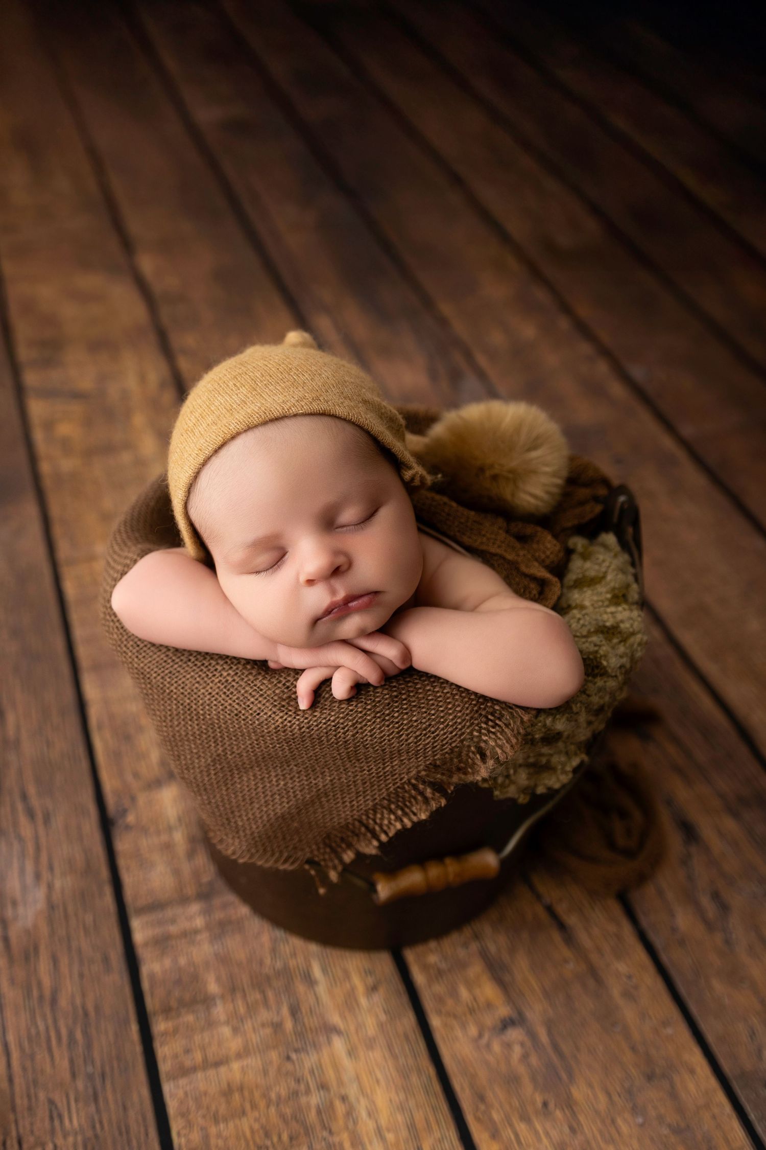 A newborn baby boy wearing a brown sleepy hat with a pom while placed in a brown bucket