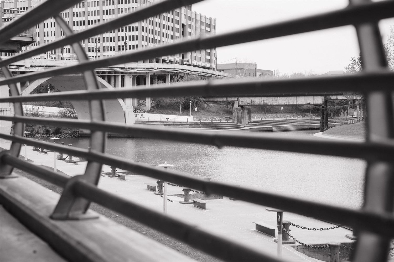 Black and white view through industrial fence railings showing a city urban landscape with benches below.