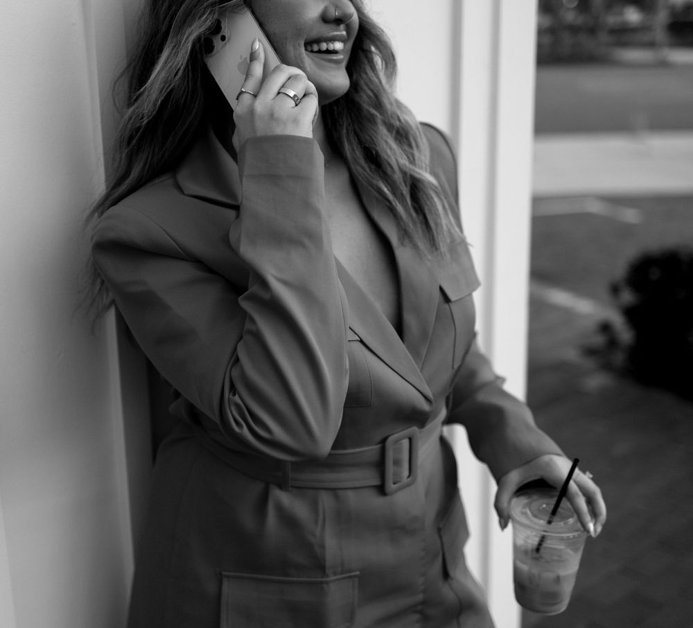 Black and white portrait of someone in a belted trench coat laughing while talking on phone.