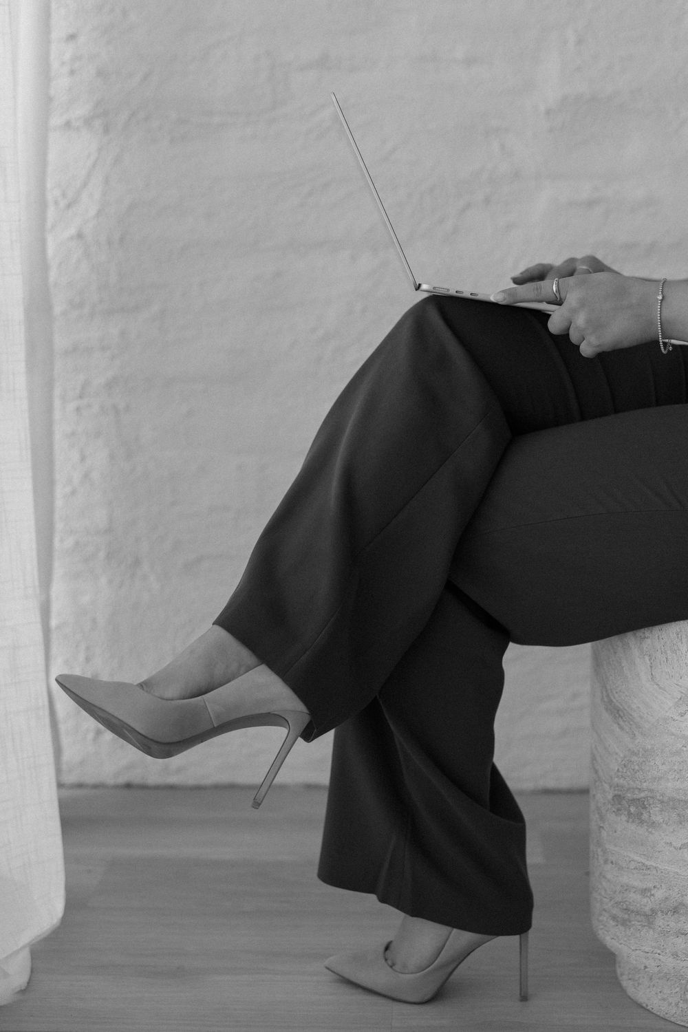 A person in black dress pants and silver stiletto heels sits casually with crossed legs against a white wall.