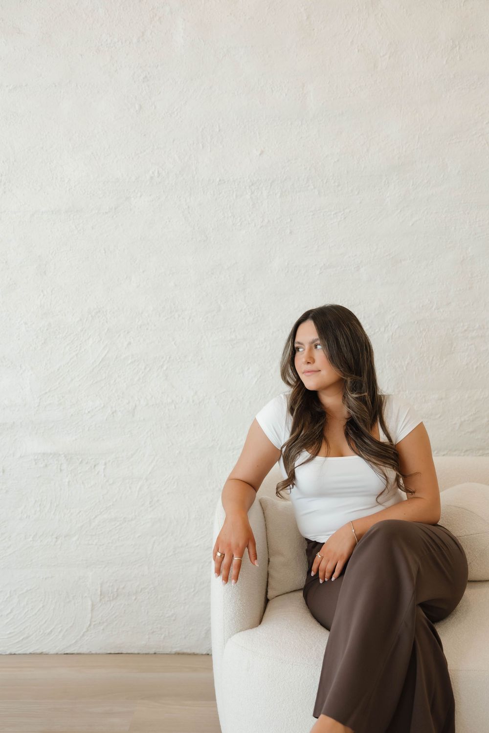 A person in a white top and brown pants sits casually on a cream colored sofa against a white textured wall.
