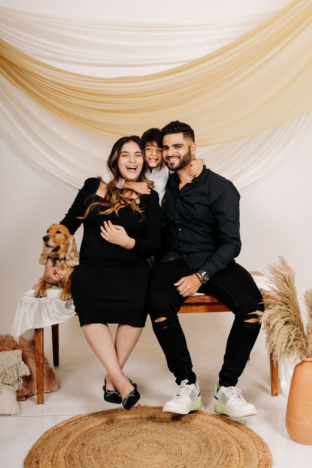 Family maternity photo with golden retriever dog against draped fabric backdrop with boho decor.