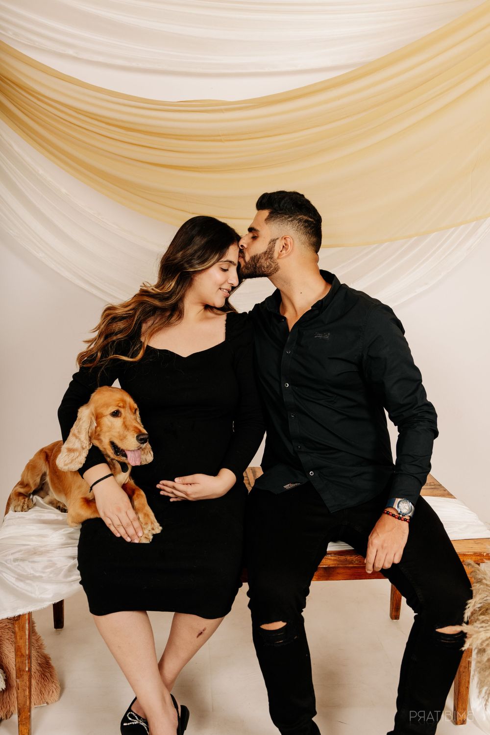 Intimate maternity photo with couple and dog against white draped backdrop.
