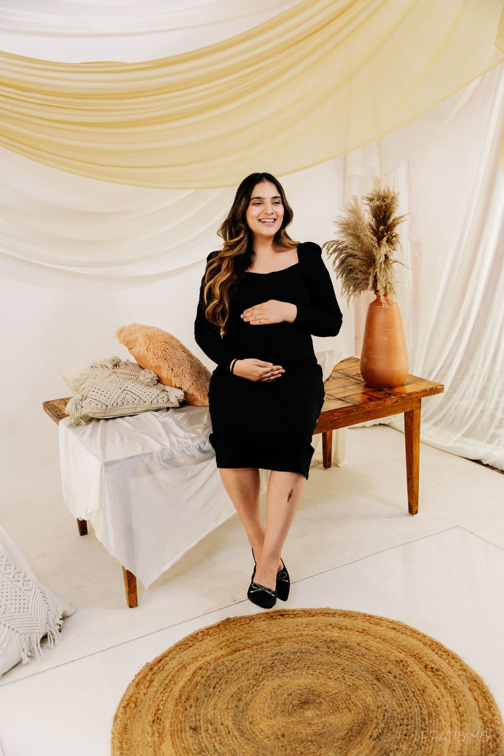 Maternity portrait of woman in black dress seated on wooden bench with boho decor.