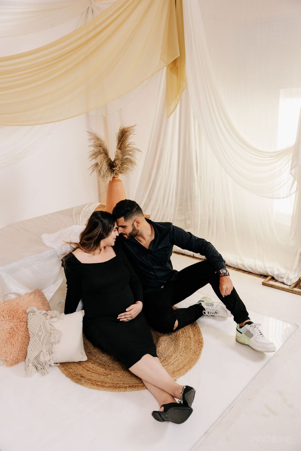 Intimate maternity photo of couple sitting on jute rug with white draped backdrop.