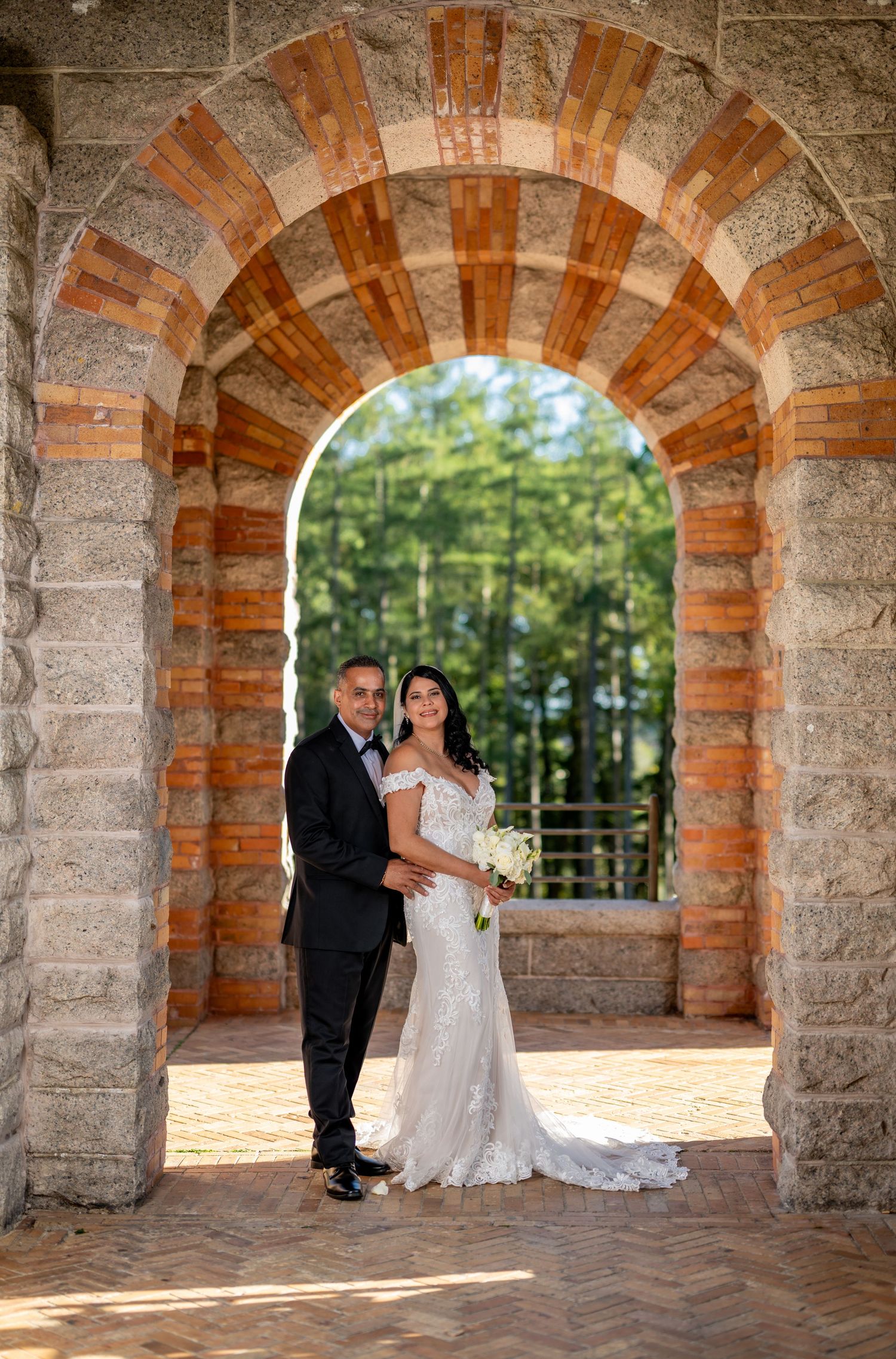 Bride and groom posing under a stone arch at their wedding venue, surrounded by lush green trees.