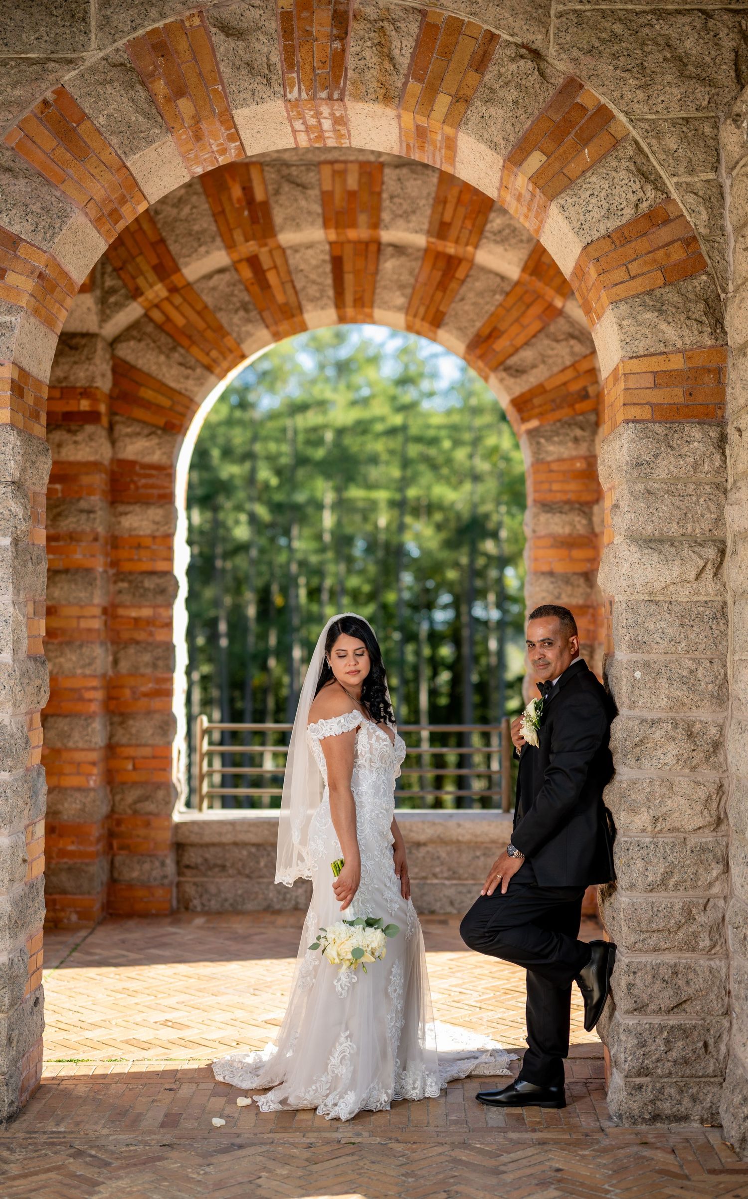 Bride in white lace gown and groom in black suit pose under a stone arch with trees in the background.