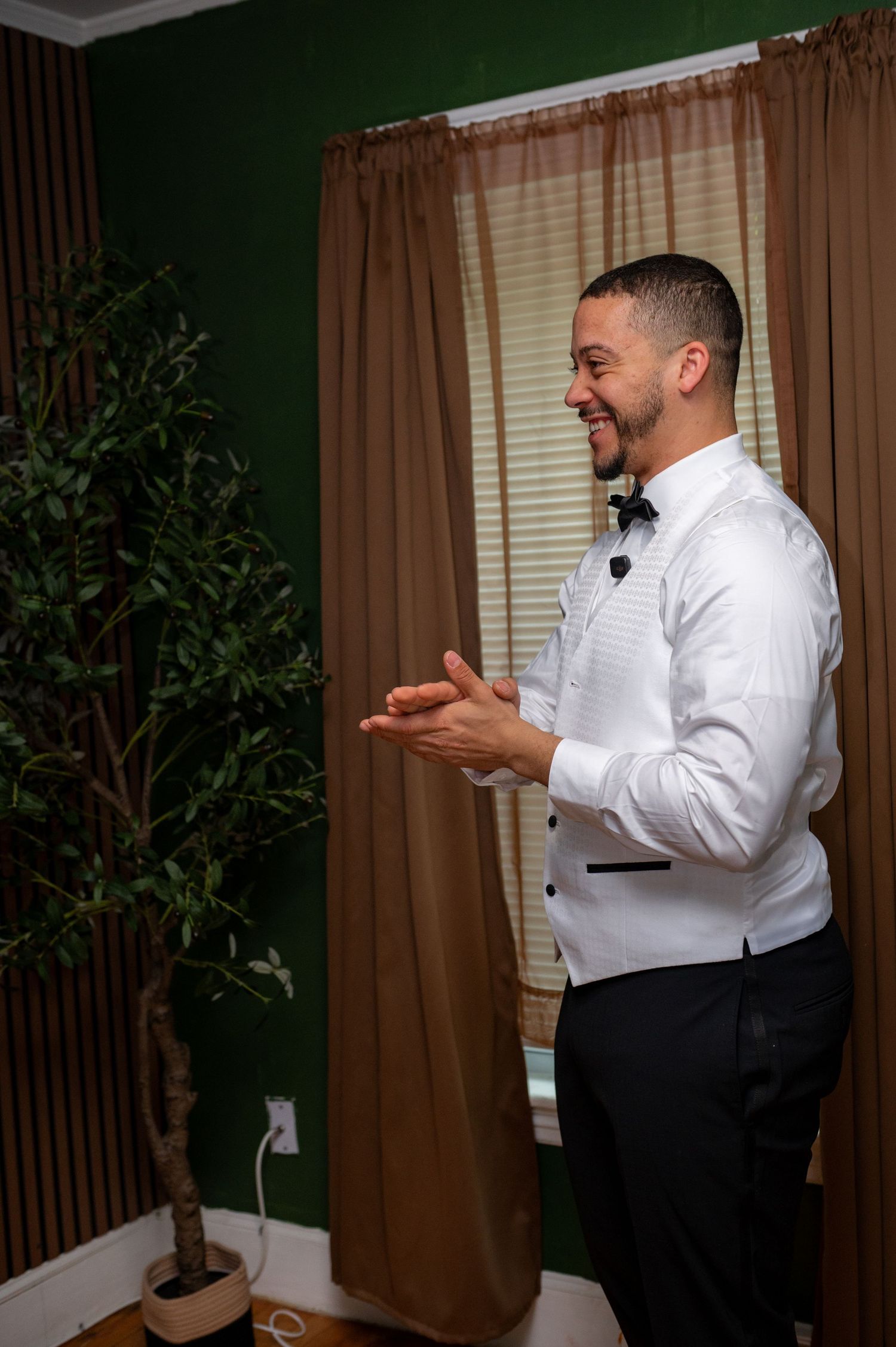Smiling groom in white tuxedo shirt and bow tie clapping hands in green decorated room before wedding ceremony.