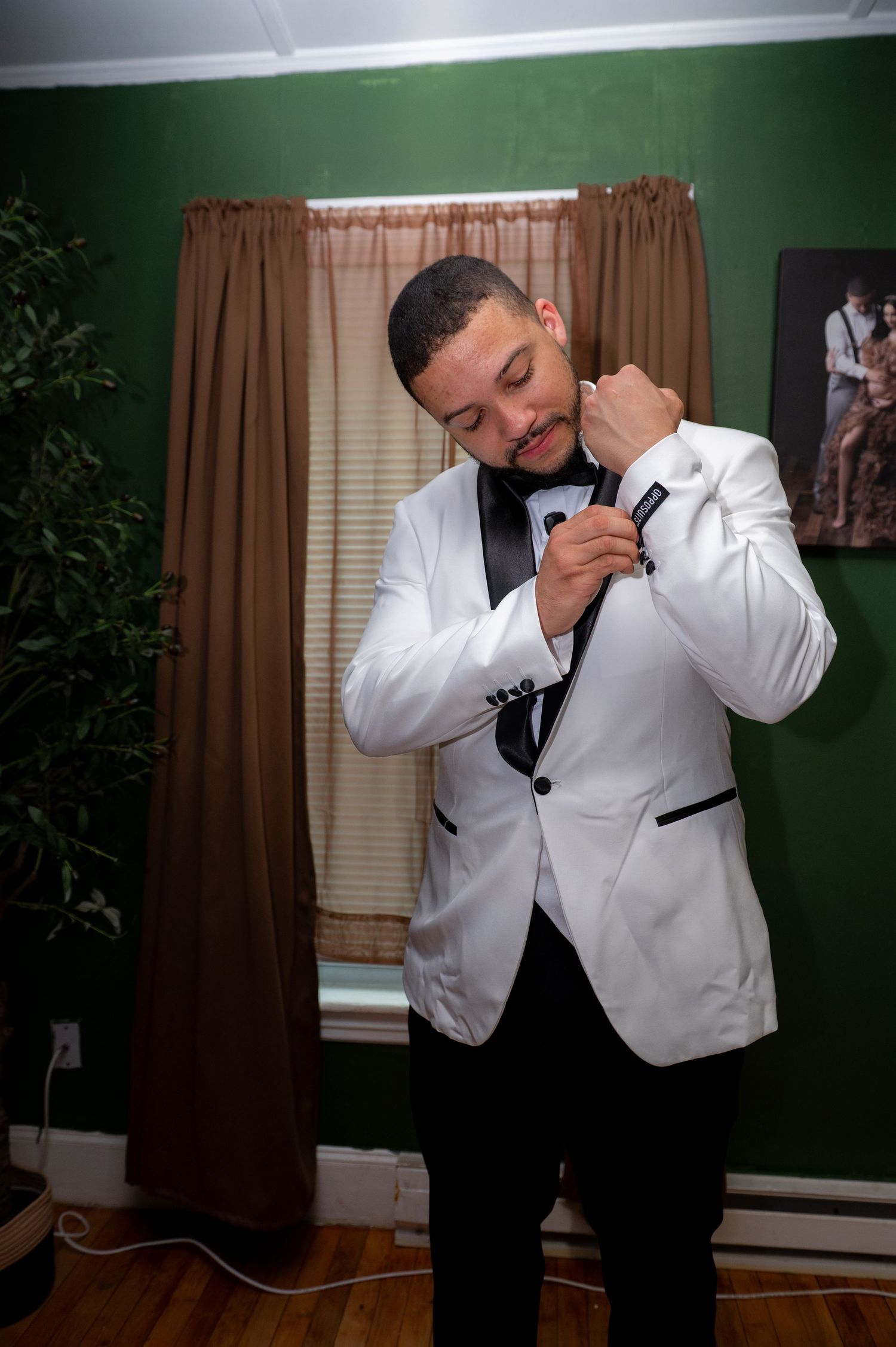 A young man in a white tuxedo jacket adjusts his bow tie while getting dressed in a green-walled room.