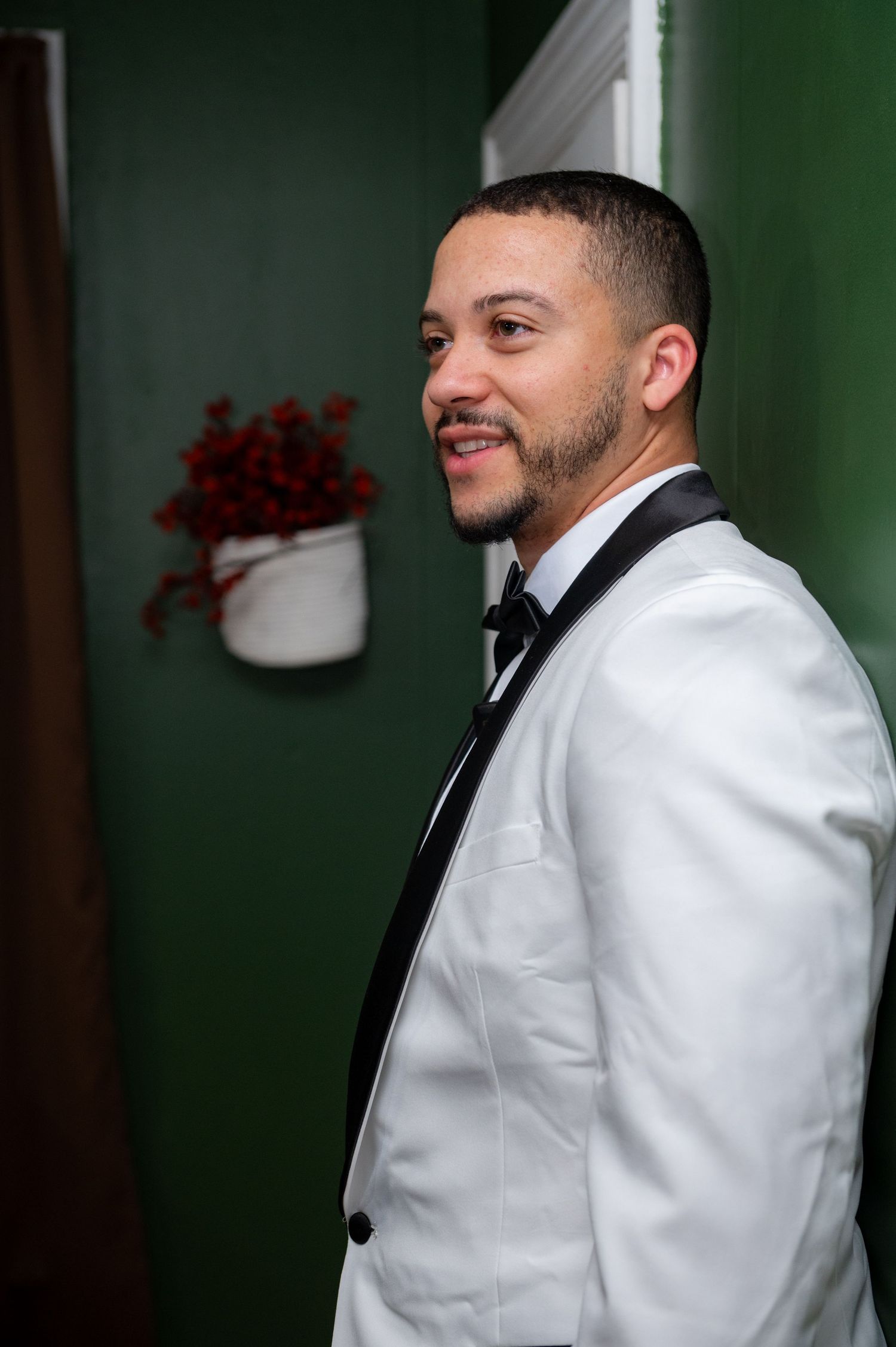 Man in white tuxedo jacket and black bow tie smiling, standing near green wall with red floral decor.