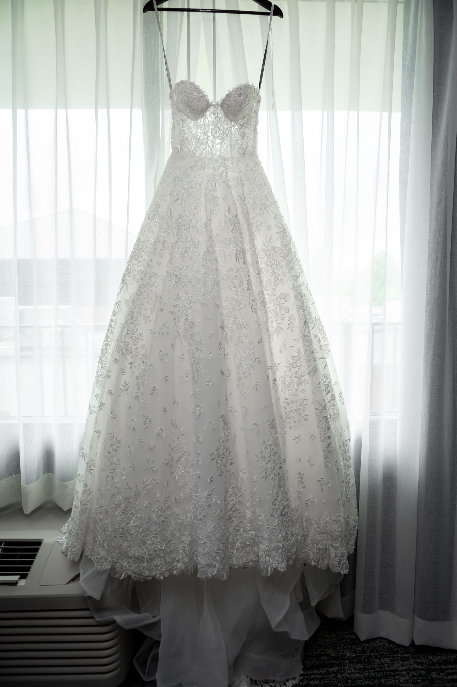 White lace ball gown wedding dress with sweetheart neckline hanging in front of sheer curtained window.