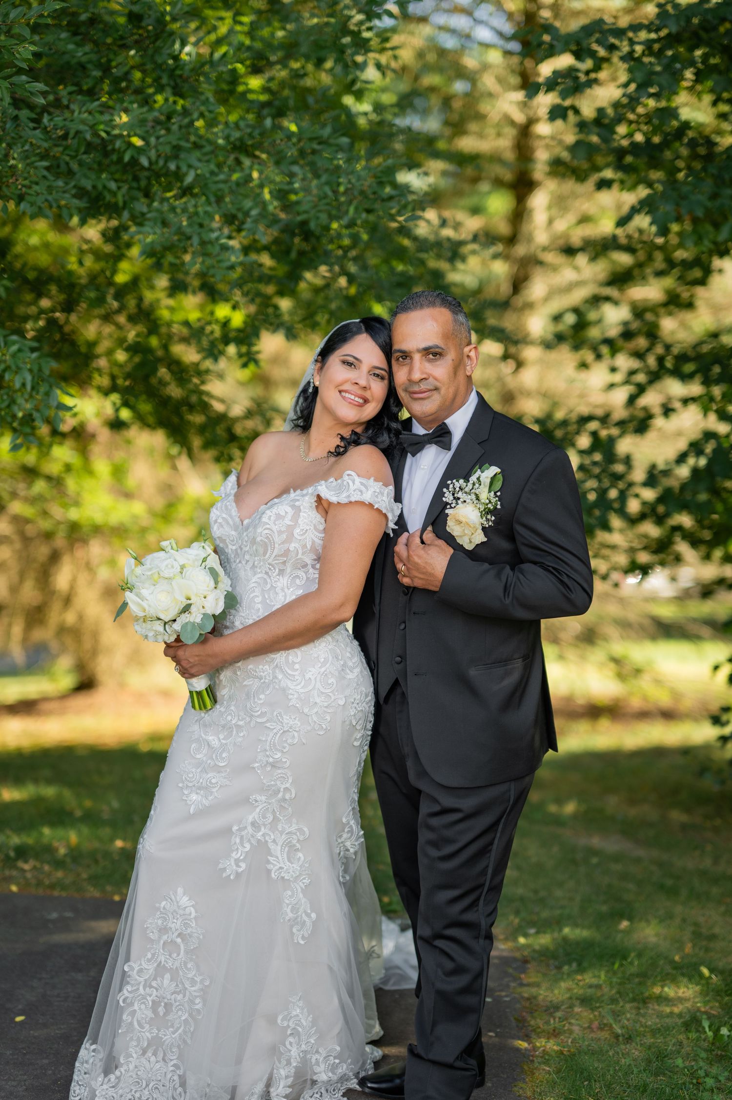 Bride in lace off-shoulder gown holding white bouquet poses with groom in black tuxedo outdoors.