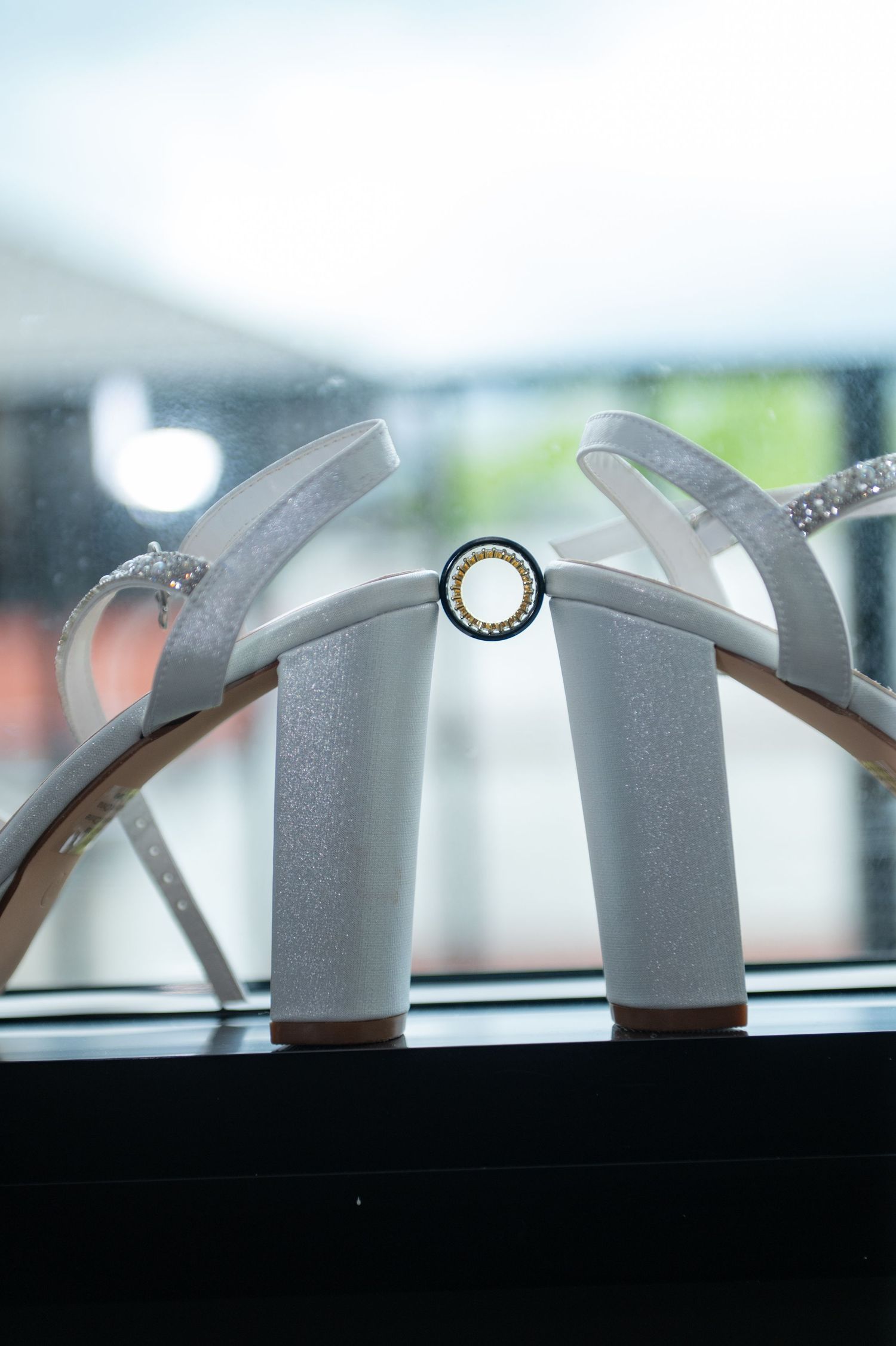 White glitter block heel wedding sandals displayed on a windowsill with a small decorative brooch between the heels.