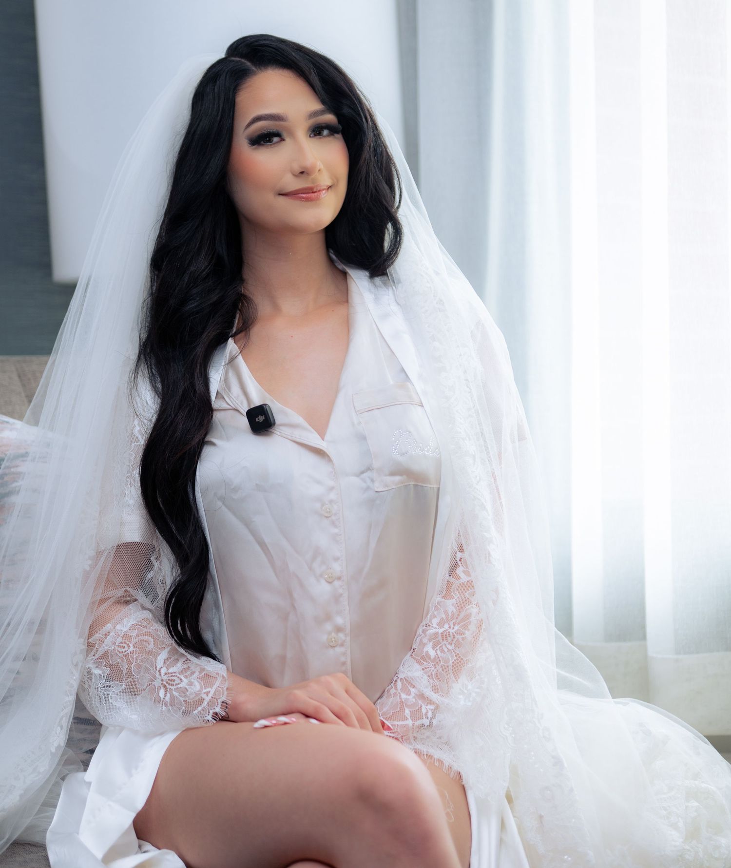 A bride in a white lace robe and veil with long dark hair poses elegantly in a bright, airy room.