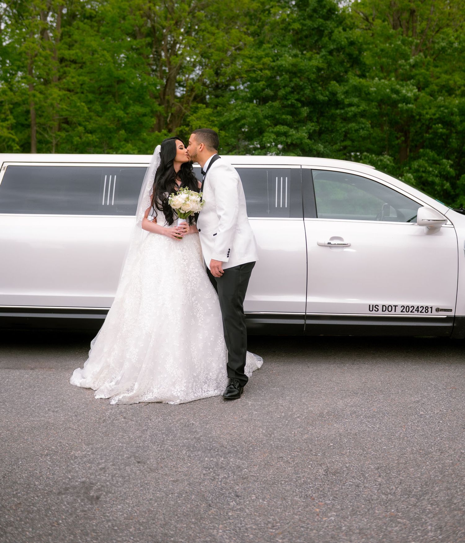 Newlywed couple kissing in front of a white stretch limousine on their wedding day, bride in lace gown holding bouquet.