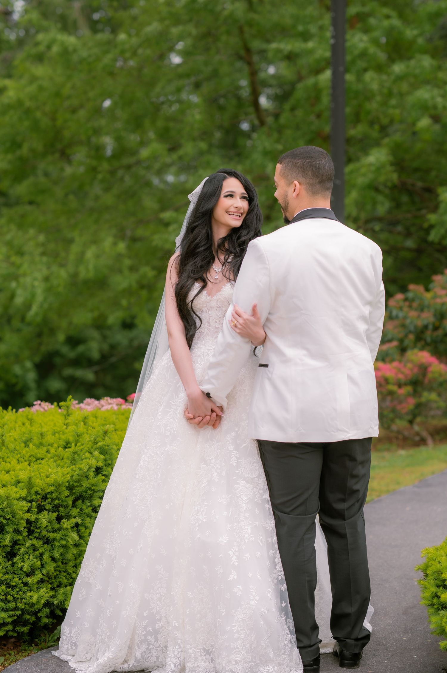 Bride in lace gown and veil smiles at groom in white tuxedo jacket while holding hands in a garden.