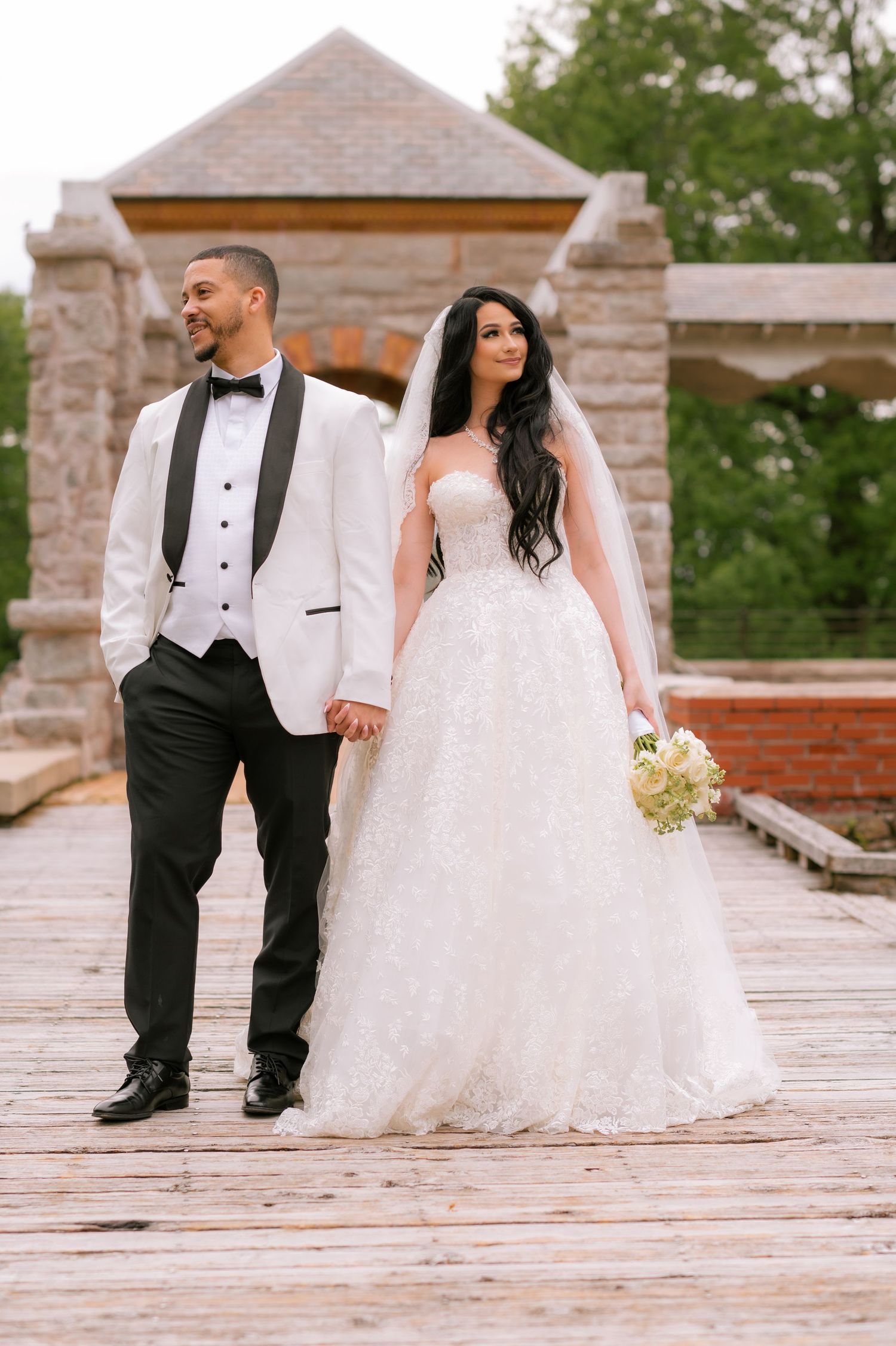 Bride in white ball gown and groom in white tuxedo holding hands on wooden deck before stone pavilion.