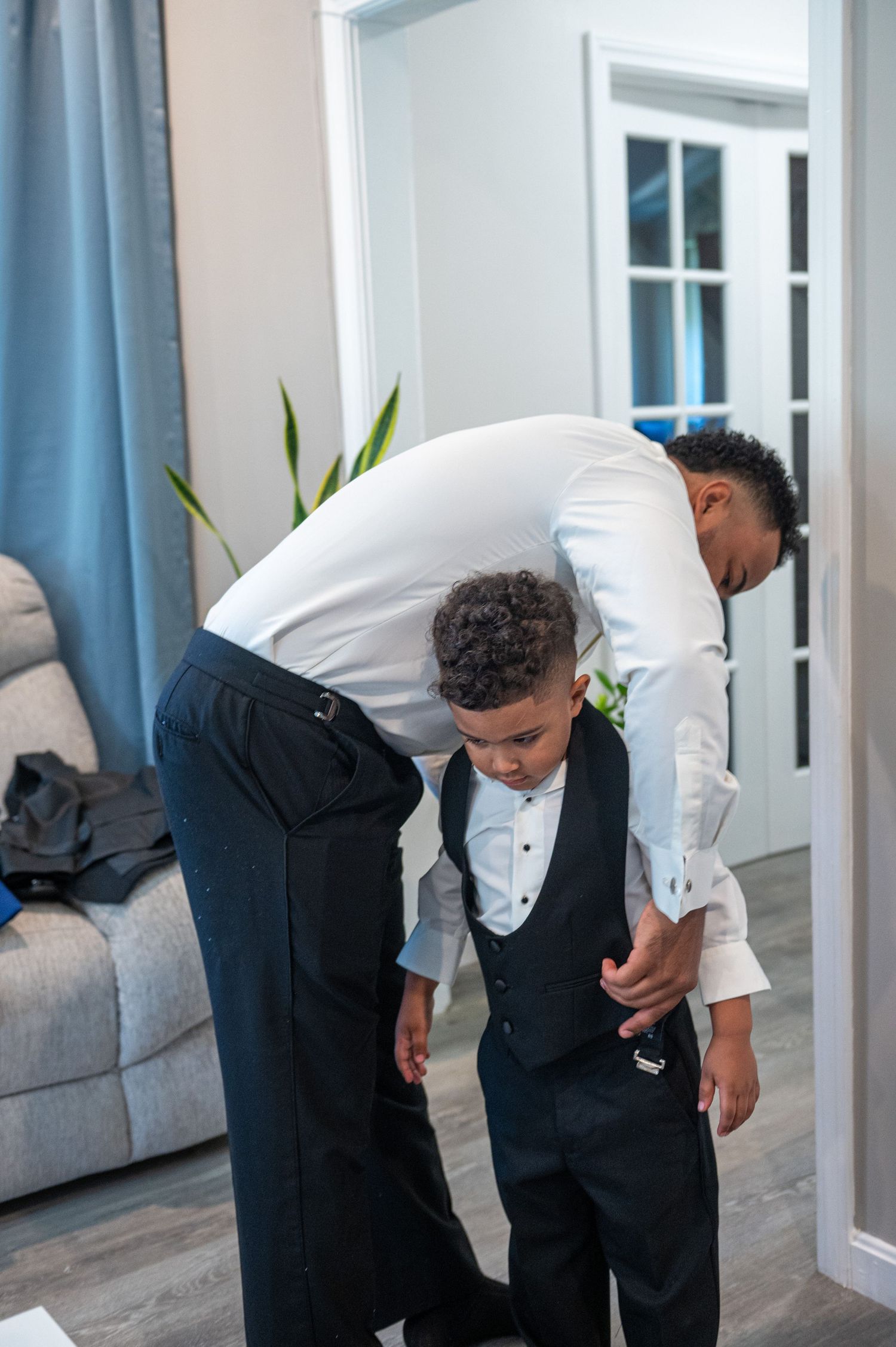 Two people in formal attire helping with wedding day preparations in a living room.
