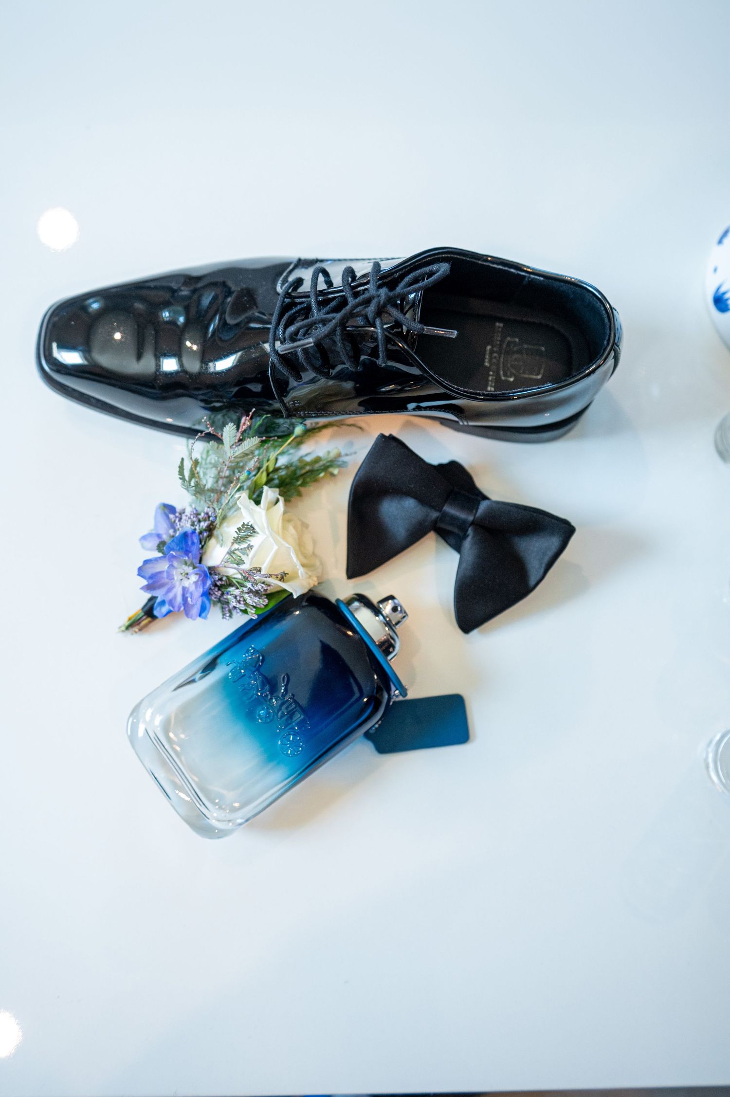 Wedding accessories including dress shoes, bow tie, and blue cologne bottle with floral decor.