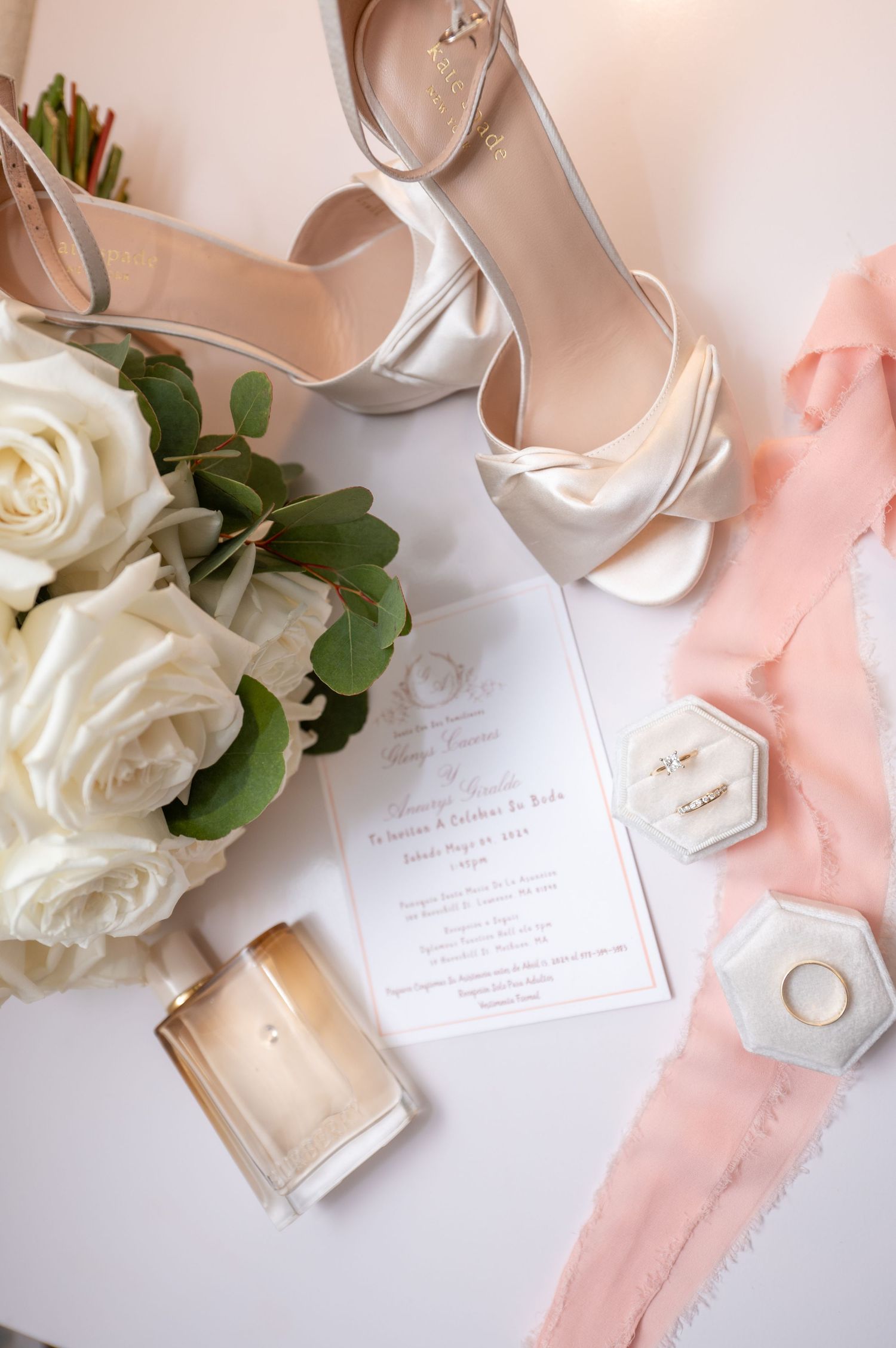 Wedding accessories flatlay with white roses, silver shoes, and blush pink ribbon on light background.