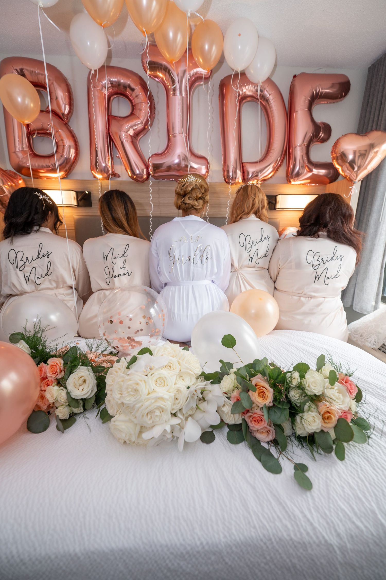 Bridal party robes and rose gold balloon decorations with floral arrangements on white bed.