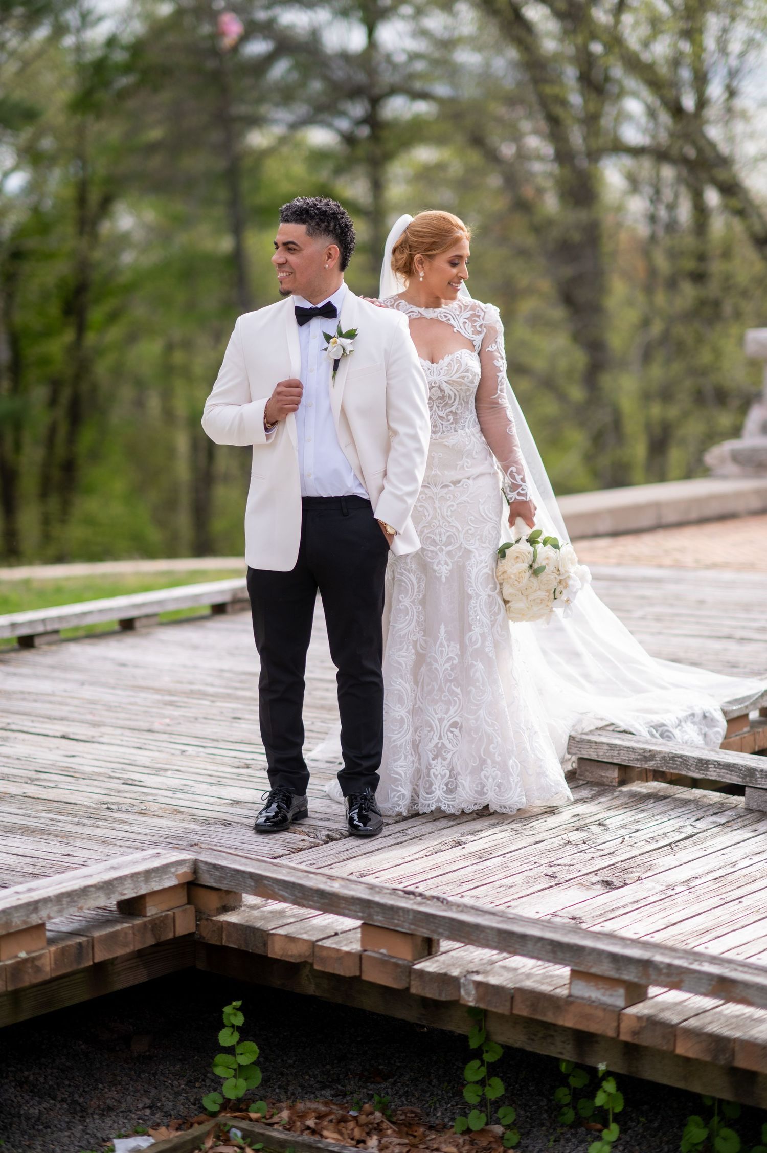 Wedding couple walks on wooden deck surrounded by spring greenery.