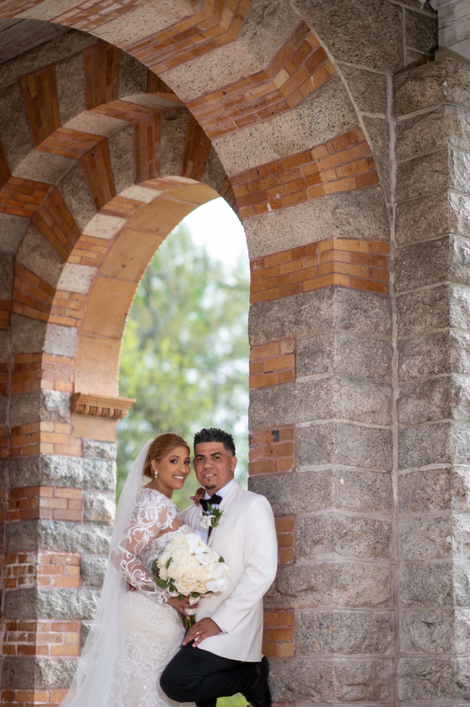 A romantic wedding moment captured under a beautiful brick and stone archway with a floral bouquet.