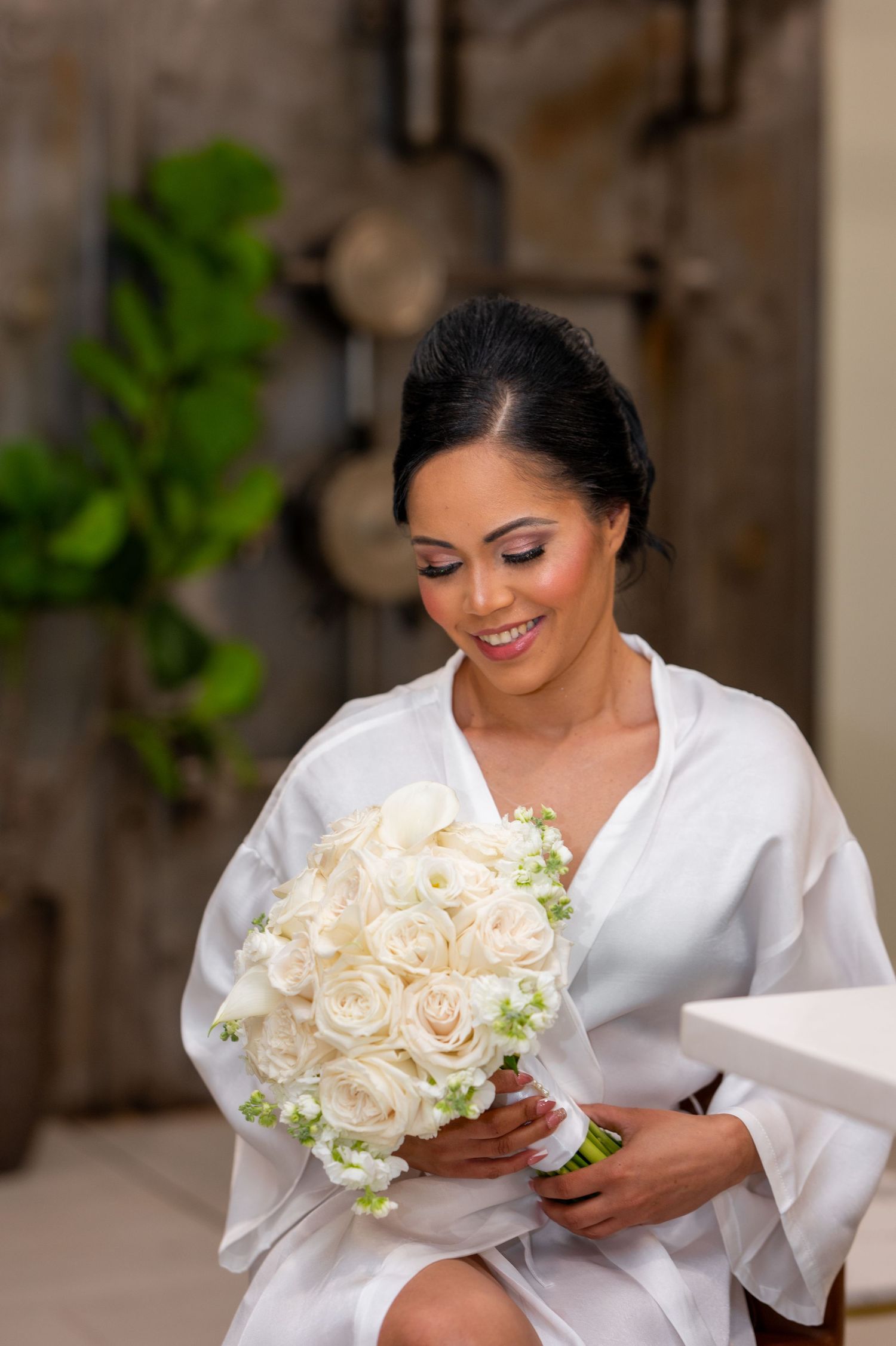 Smiling bride in white robe holding elegant bouquet of white roses and flowers, looking down with soft makeup.