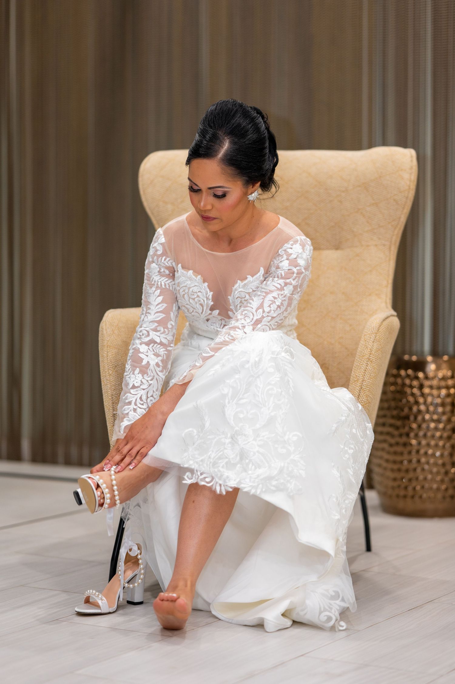 Bride in lace long-sleeve wedding gown sitting in chair, fastening pearl ankle strap heels before ceremony.