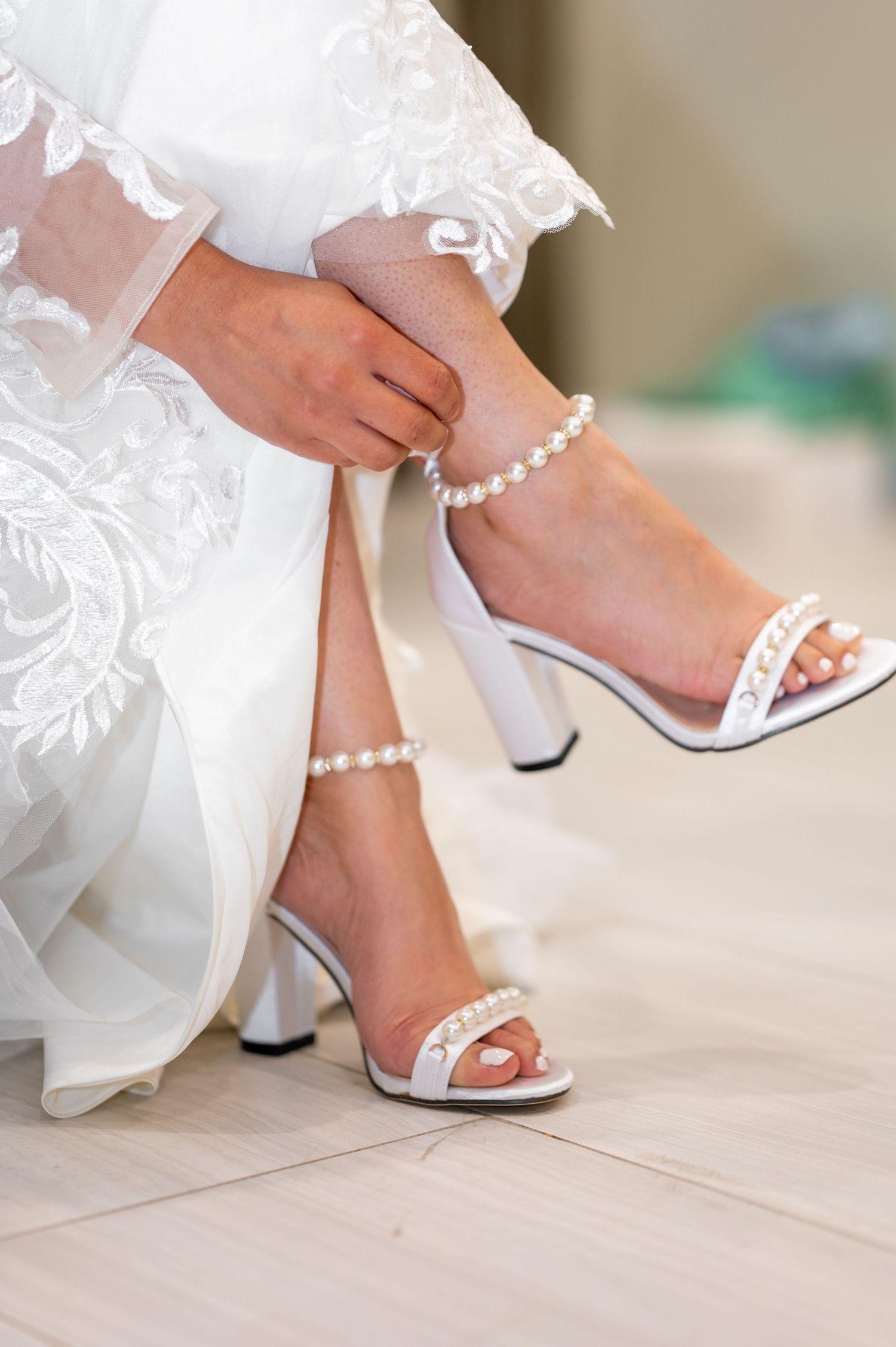Bride adjusting pearl-adorned white block heel sandals with ankle strap, wearing lace wedding gown.