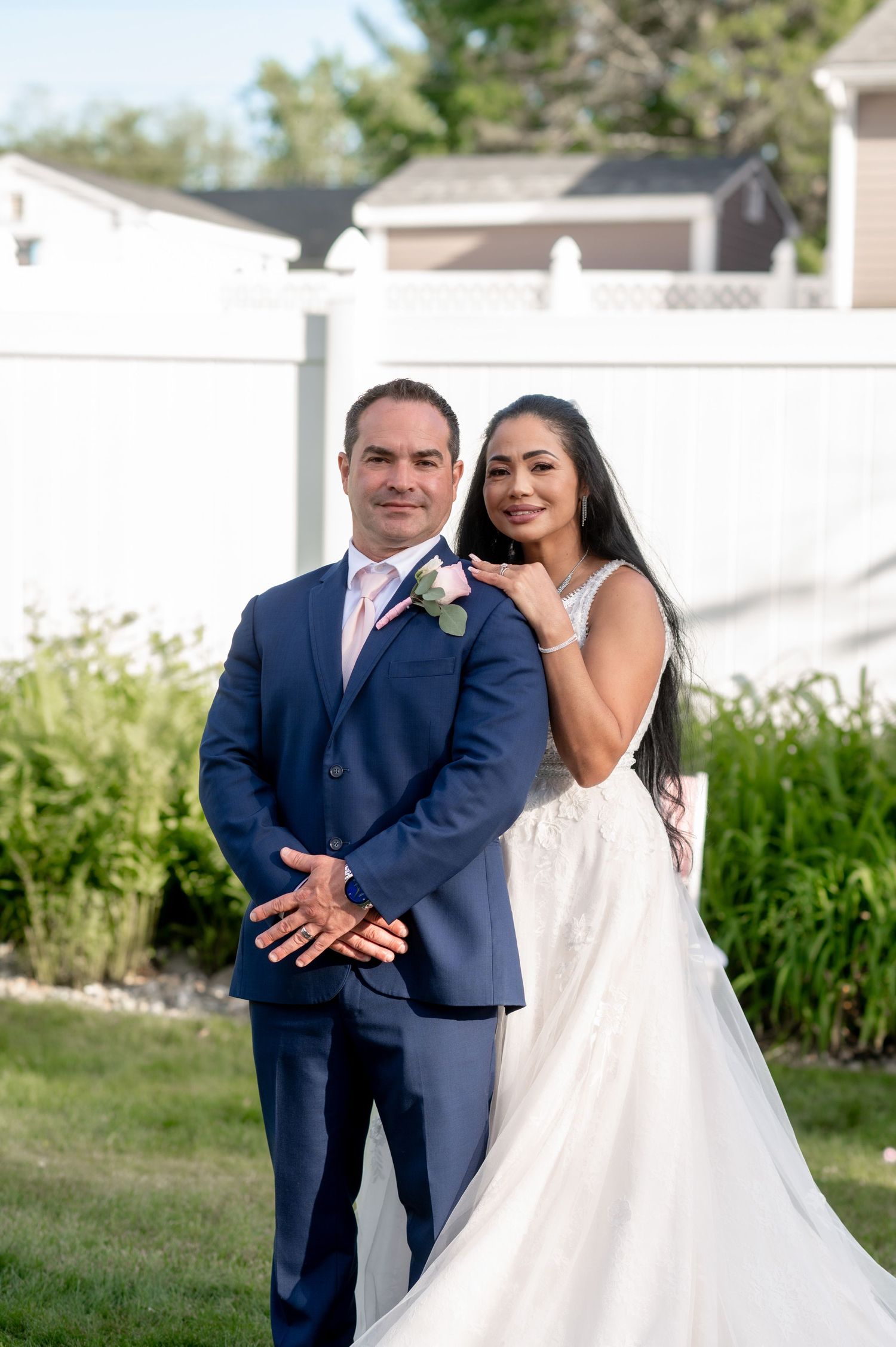 A happy wedding couple poses in front of a white fence during their outdoor celebration.