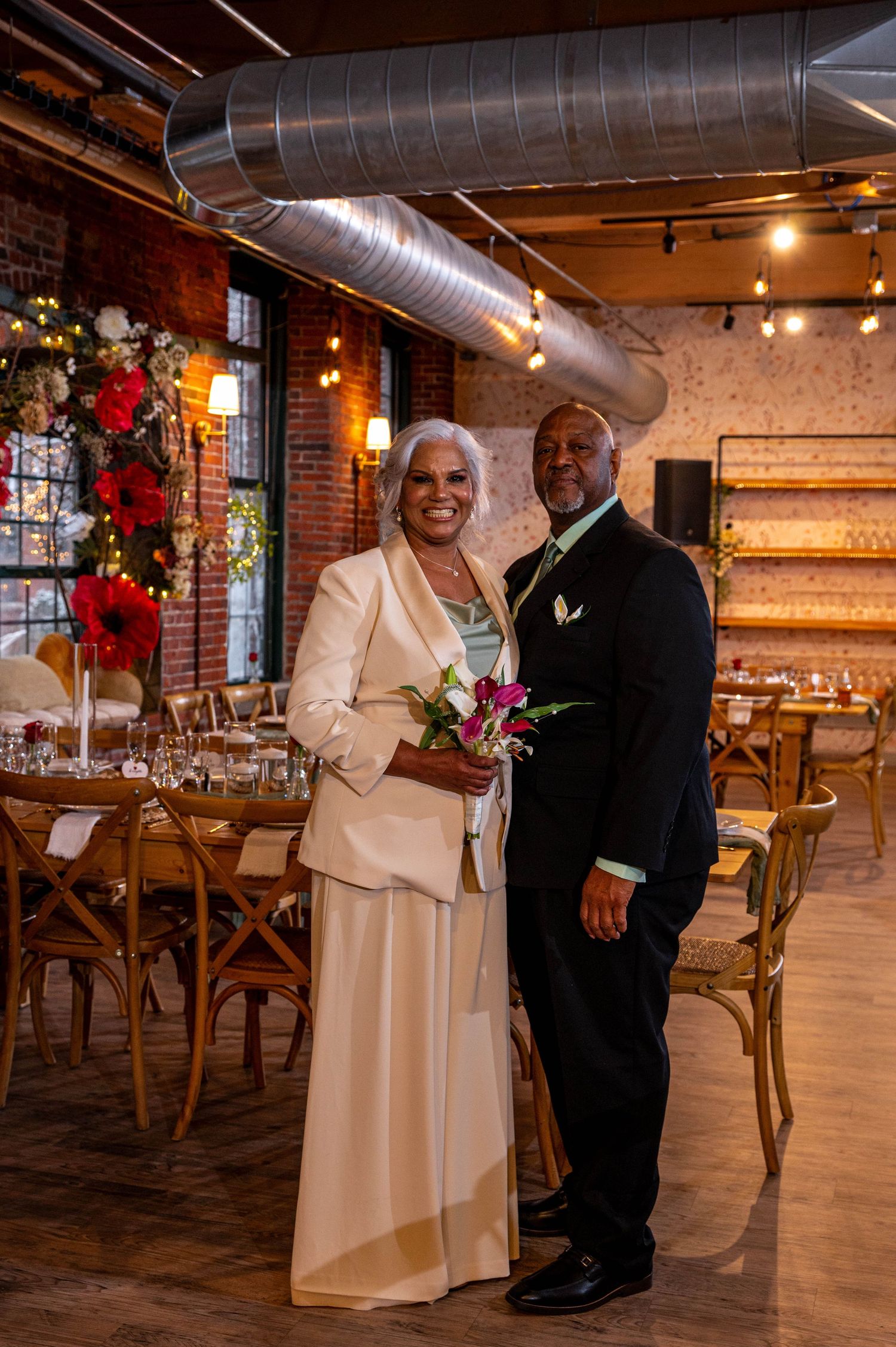 Elegant couple posing in a rustic wedding venue with brick walls, string lights, and floral arrangements.
