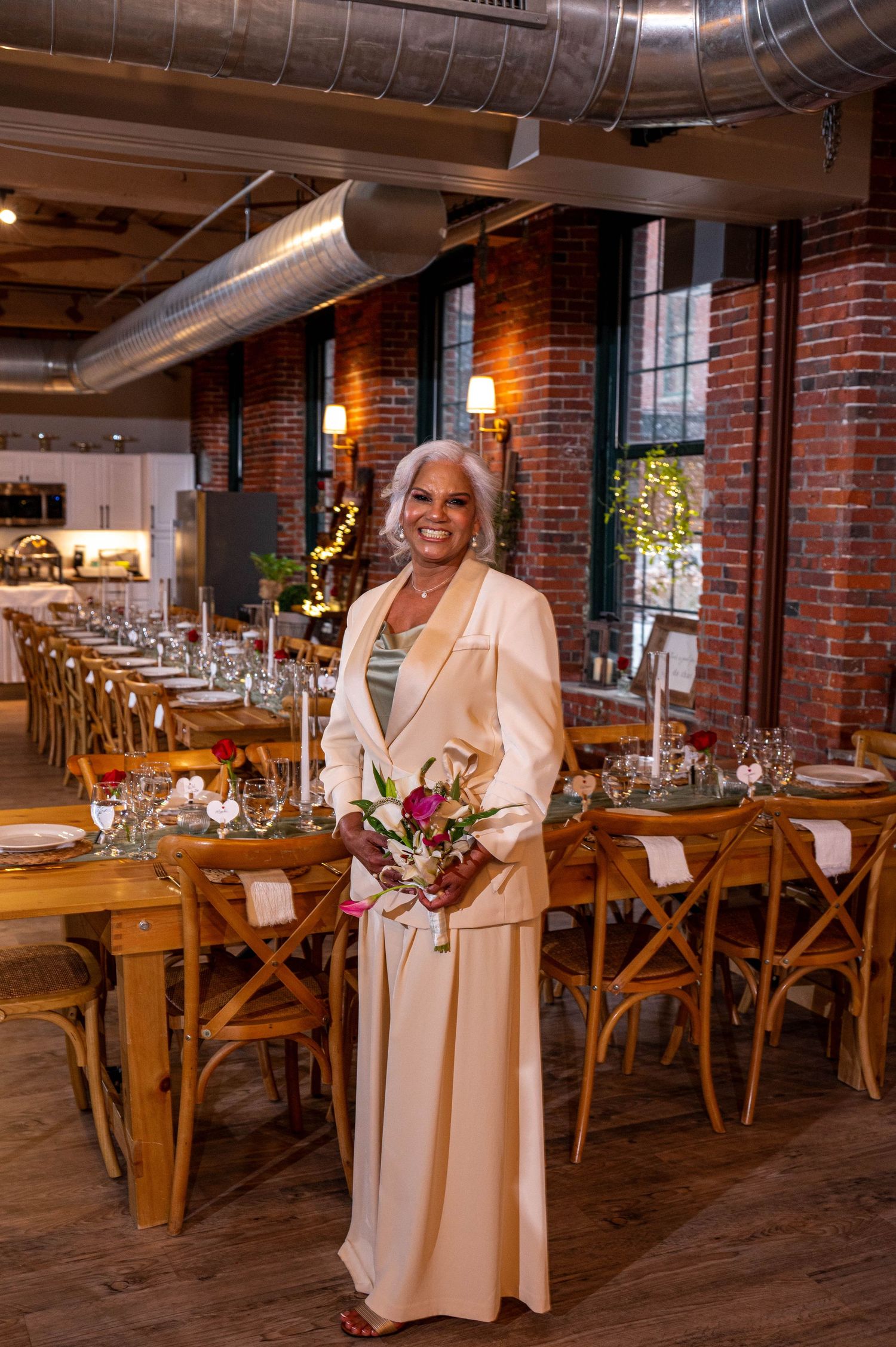 Elegant woman in cream suit holding bouquet stands in rustic brick wedding venue with long decorated tables.