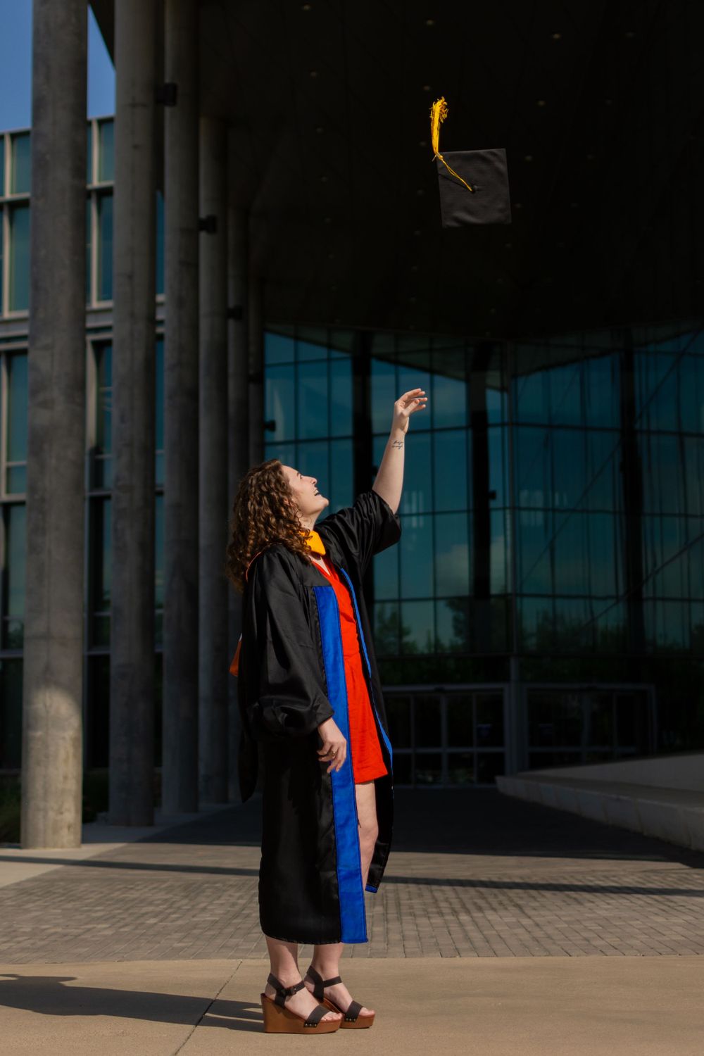 UTA graduate student stands & tosses graduation cap in the air, tassel flowing. She wears her gown & academic hood with orange dress peeking