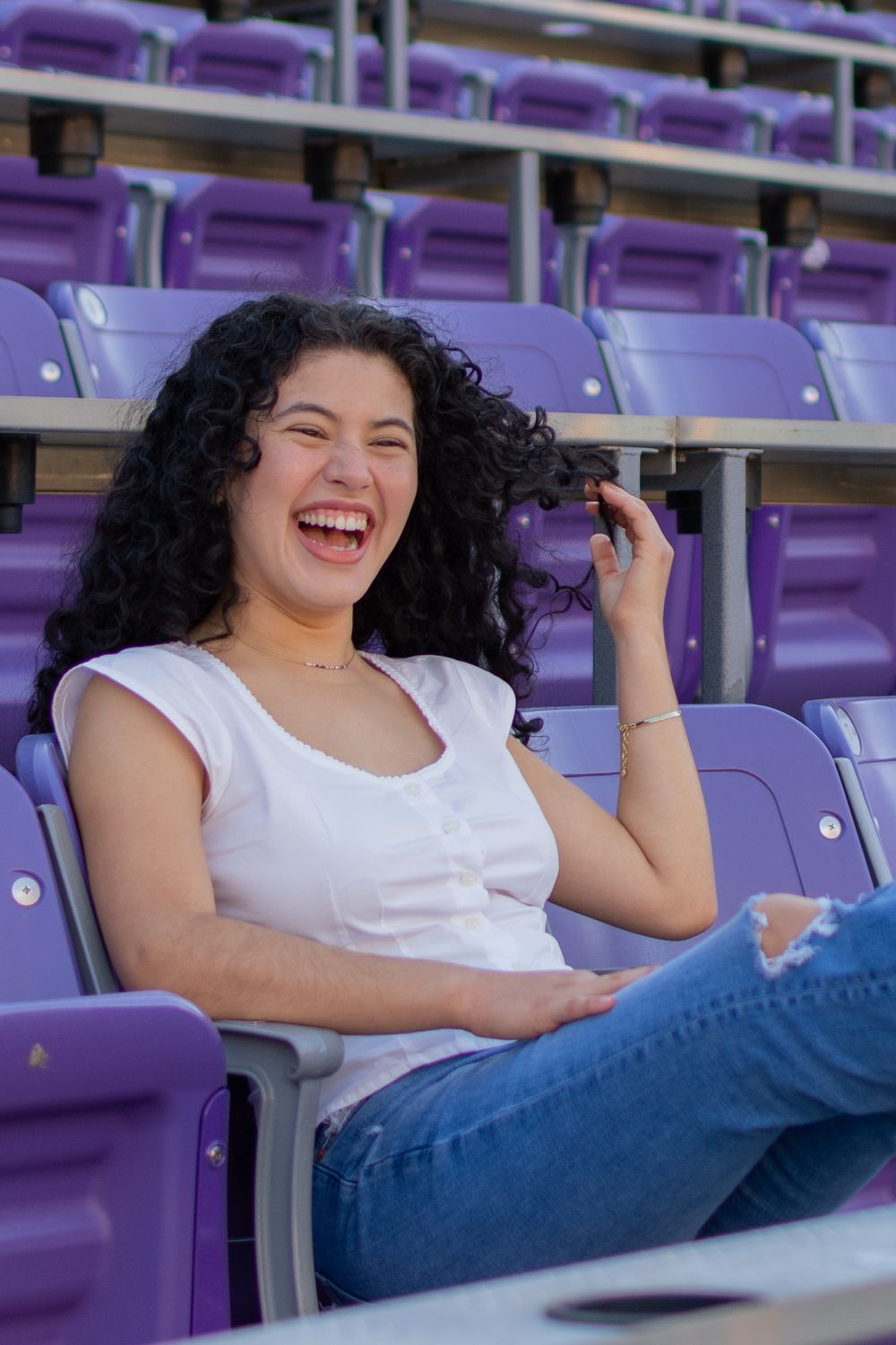 A TCU grad sits in purple, horned frogs football stadium seats, laughing and playing with her hair. She wears a white top and ripped jeans.