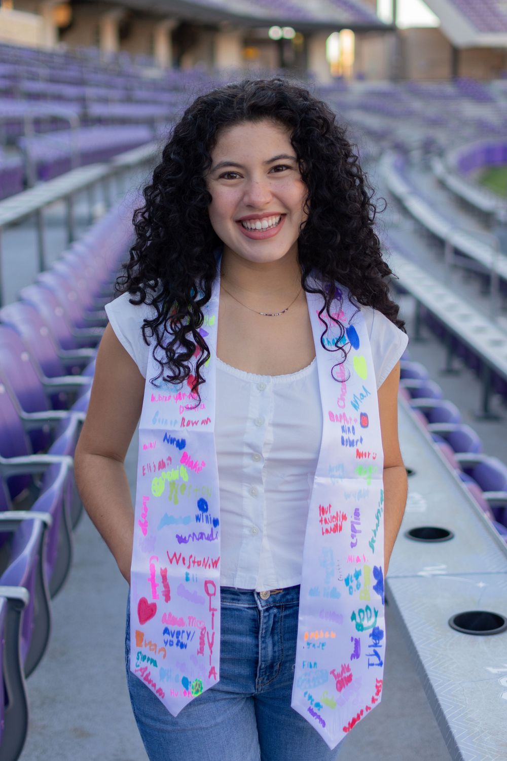 A TCU grad stands and smiles among purple, horned frogs stadium seats. She's wearing a white top, ripped jeans, & a stole signed by students