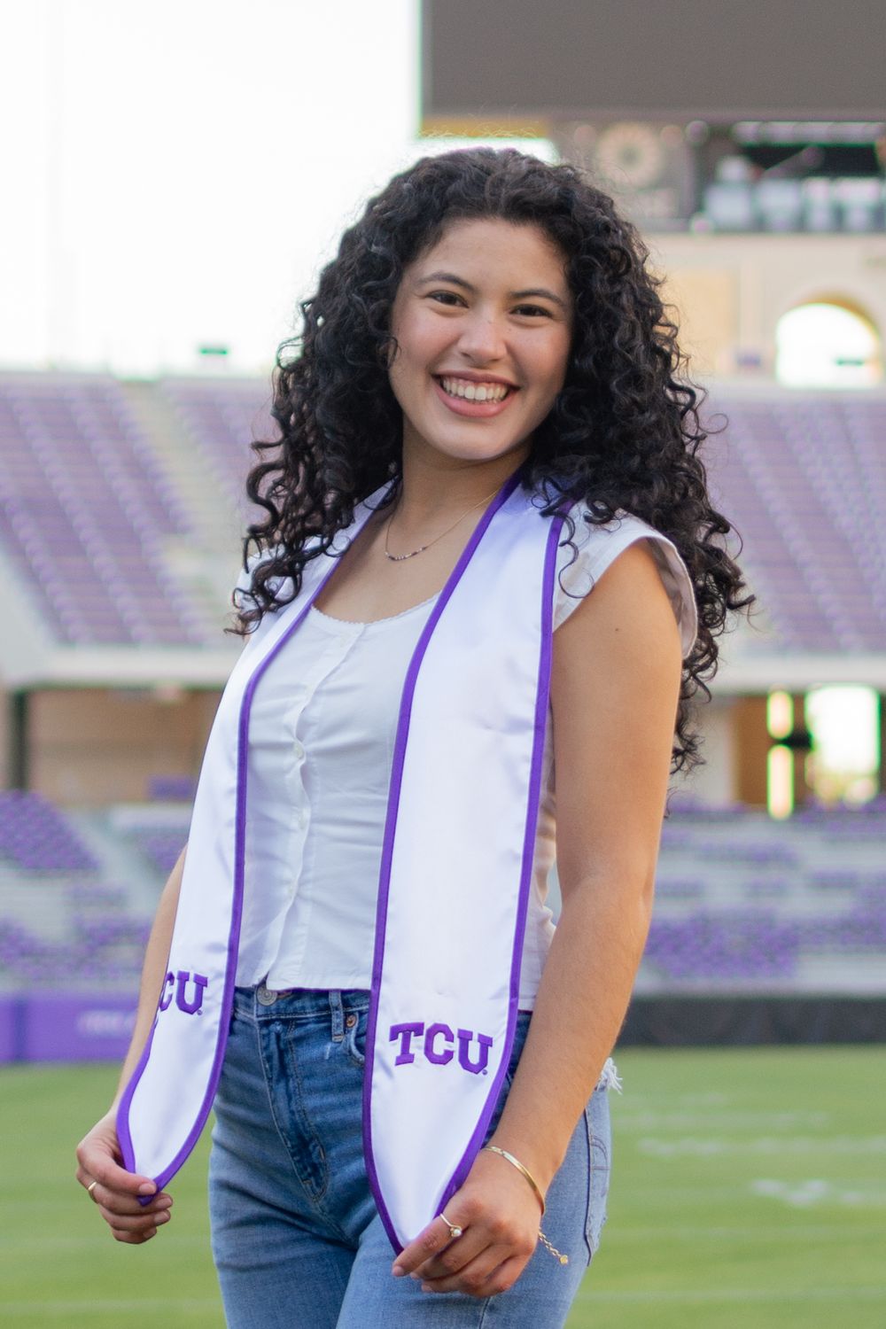 A TCU grad smiles and poses in the horned frogs football field while holding the bottom ends of her white and purple graduation stole.