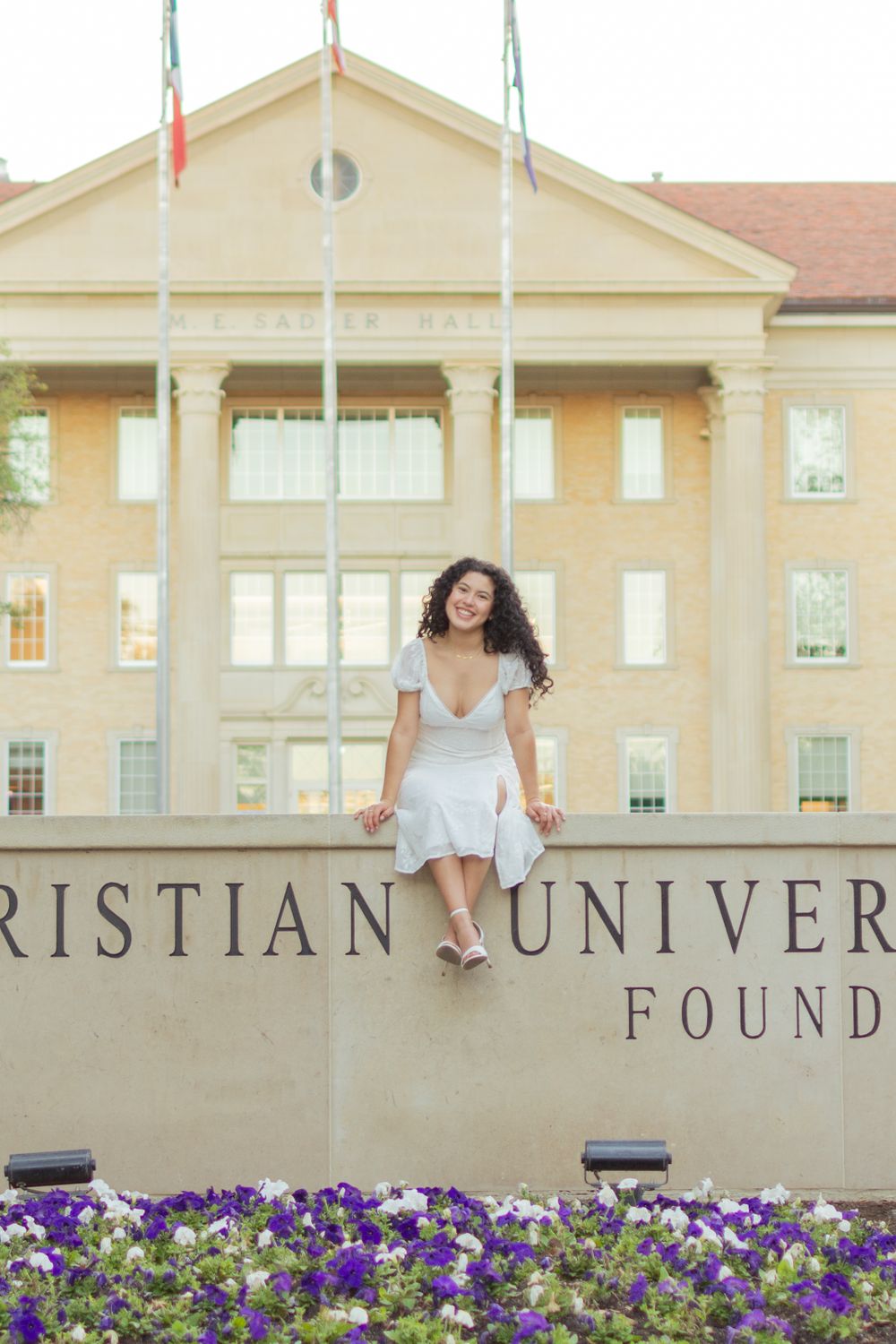 A TCU senior sits on the campus sign, leaning forward, and smiling. She's wearing a white sundress with white heels.