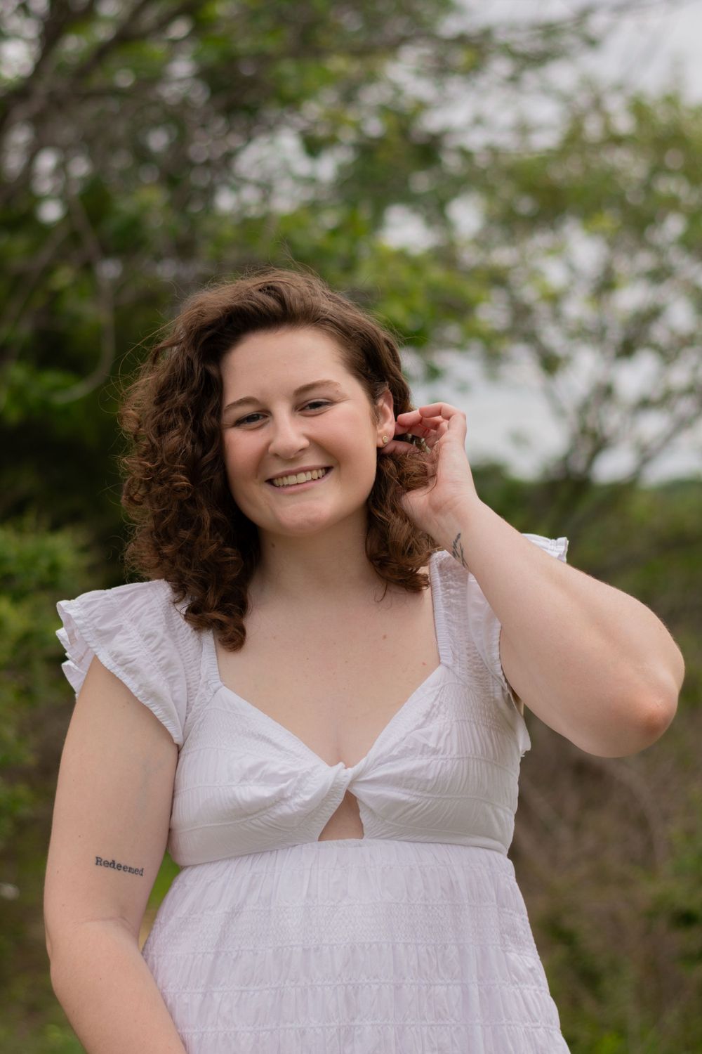 A grad tucks her hair behind one ear and smiles in her white dress during a grad photo session in Fort Worth, TX in the spring.