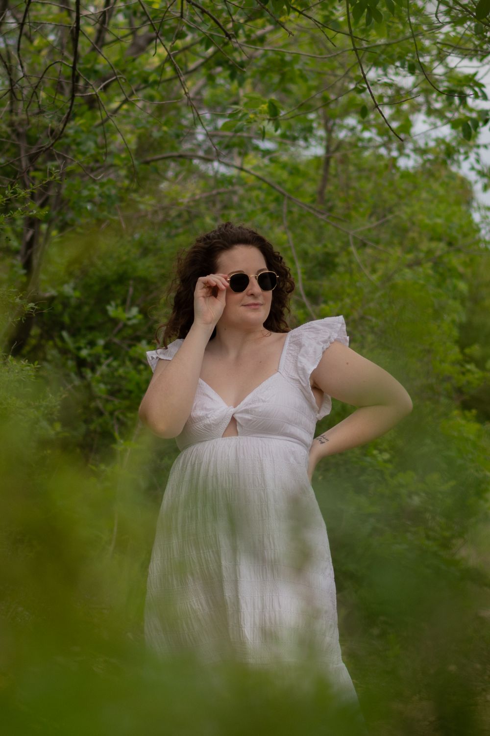 A graduate student poses in trees during her grad photos. She's wearing a white dress and sunglasses, with her right hand up to her shades.