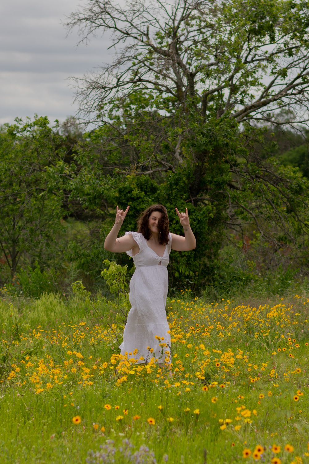 A grad holds up both hands with longhorn signs as she walks in a white dress through a flower field in Fort Worth in the spring.