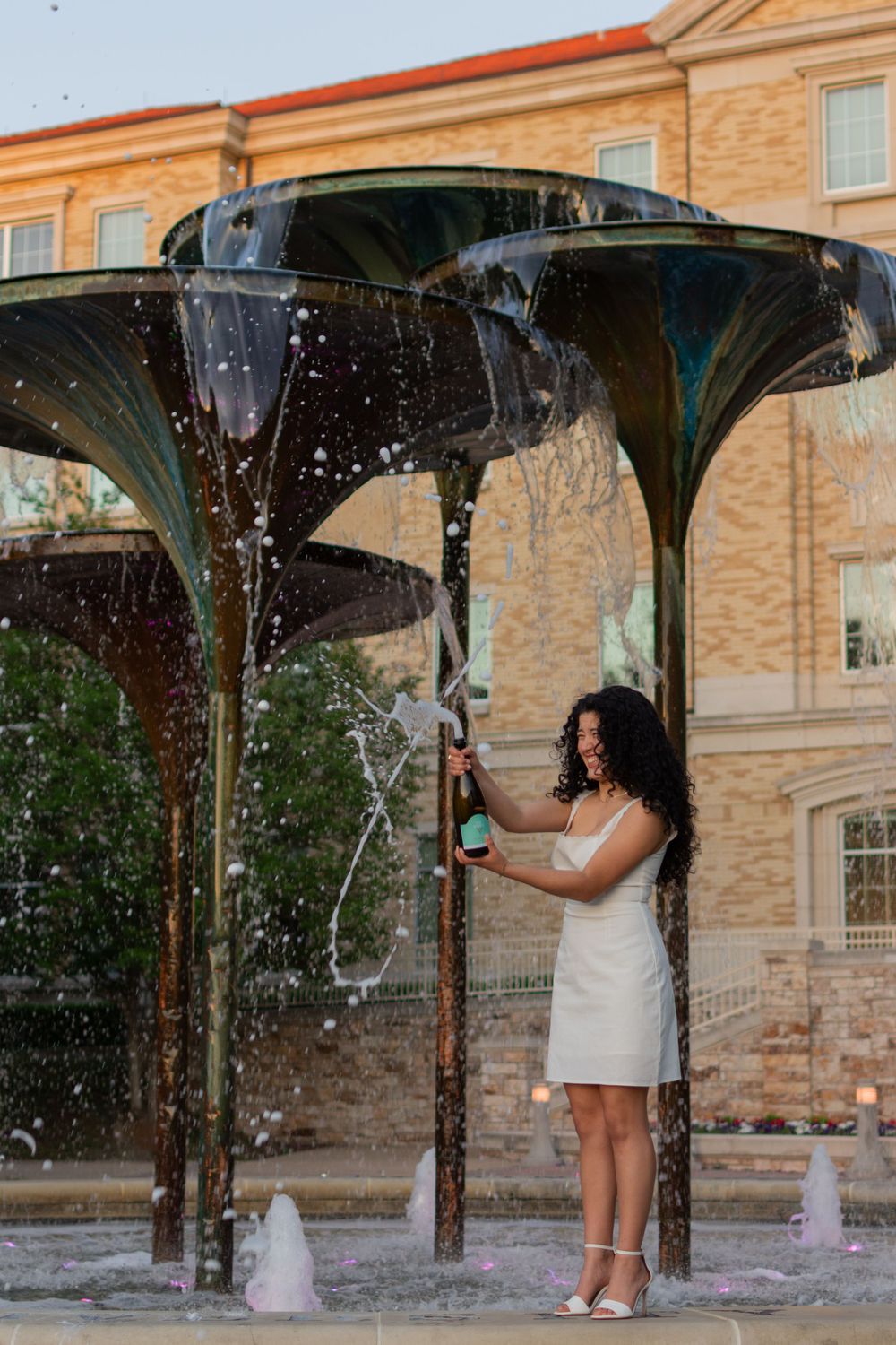 Texas Christian University grad laughs as she shakes an open bottle of champagne while standing on the edge of Frog Fountain at sunset.