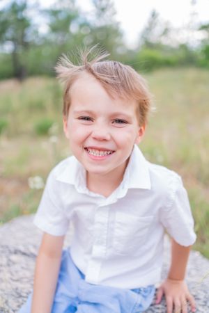 Child in white collared shirt and blue shorts sits on rock outdoors.