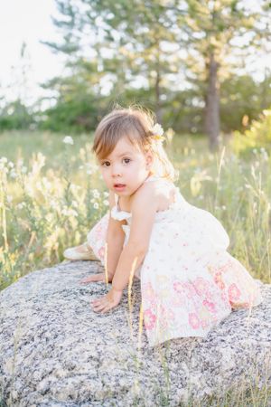 Child in floral dress sits on large rock in sunlit meadow with wildflowers.