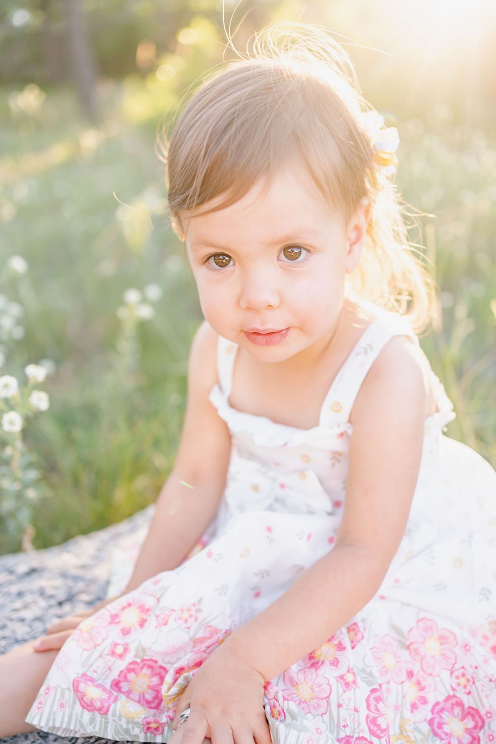 Child wearing white floral dress sits on rock during golden hour photoshoot.