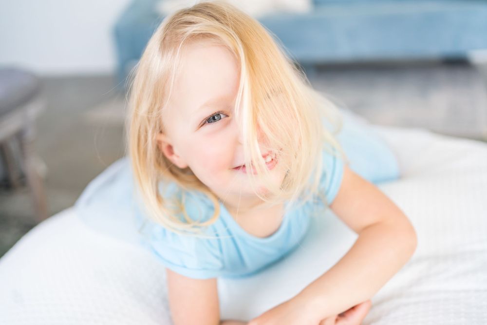 Young child with blonde hair wearing light blue dress lying on white bedding smiles at camera.