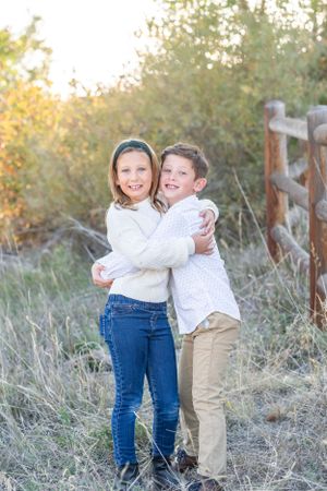 Two children embracing and laughing together in a field with fall foliage in the background.
