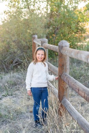 Person in white sweater and jeans stands by wooden fence in fall outdoor setting.