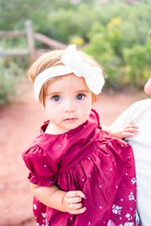 Baby in burgundy dress with white bow headband against natural backdrop.