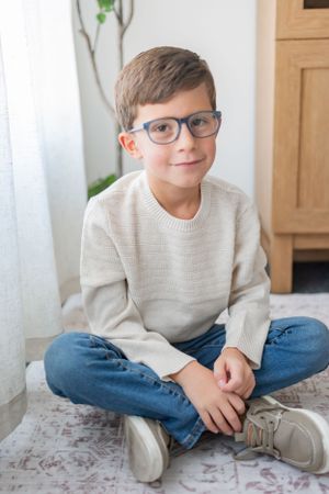 Child in cream sweater and jeans sitting cross-legged on patterned carpet.